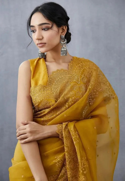 Mustard yellow organza saree with sleeveless embroidered blouse design featuring ornate lacework and grey studio backdrop