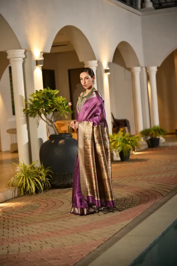 Purple tussar silk saree with golden border and ornate pallu displayed on model in traditional architectural setting with white pillars and arches